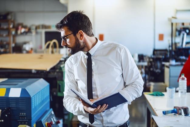 Young experienced caucasian bearded graphic engineer with eyeglasses holding notebook and checking on printing machine while standing in printing shop.