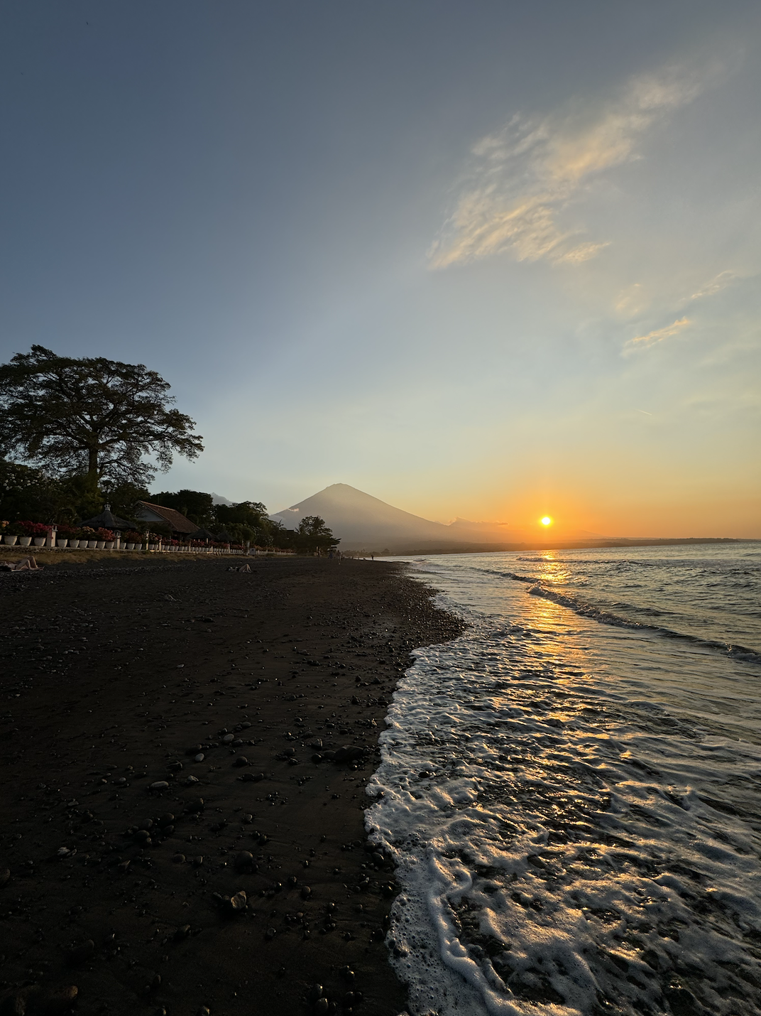 Photo of the beach at Amed, Bali. In the foreground is the waves and black sand. In the background is a sunset and the active volcano Mount Agung which erupted most recently in 2017. 