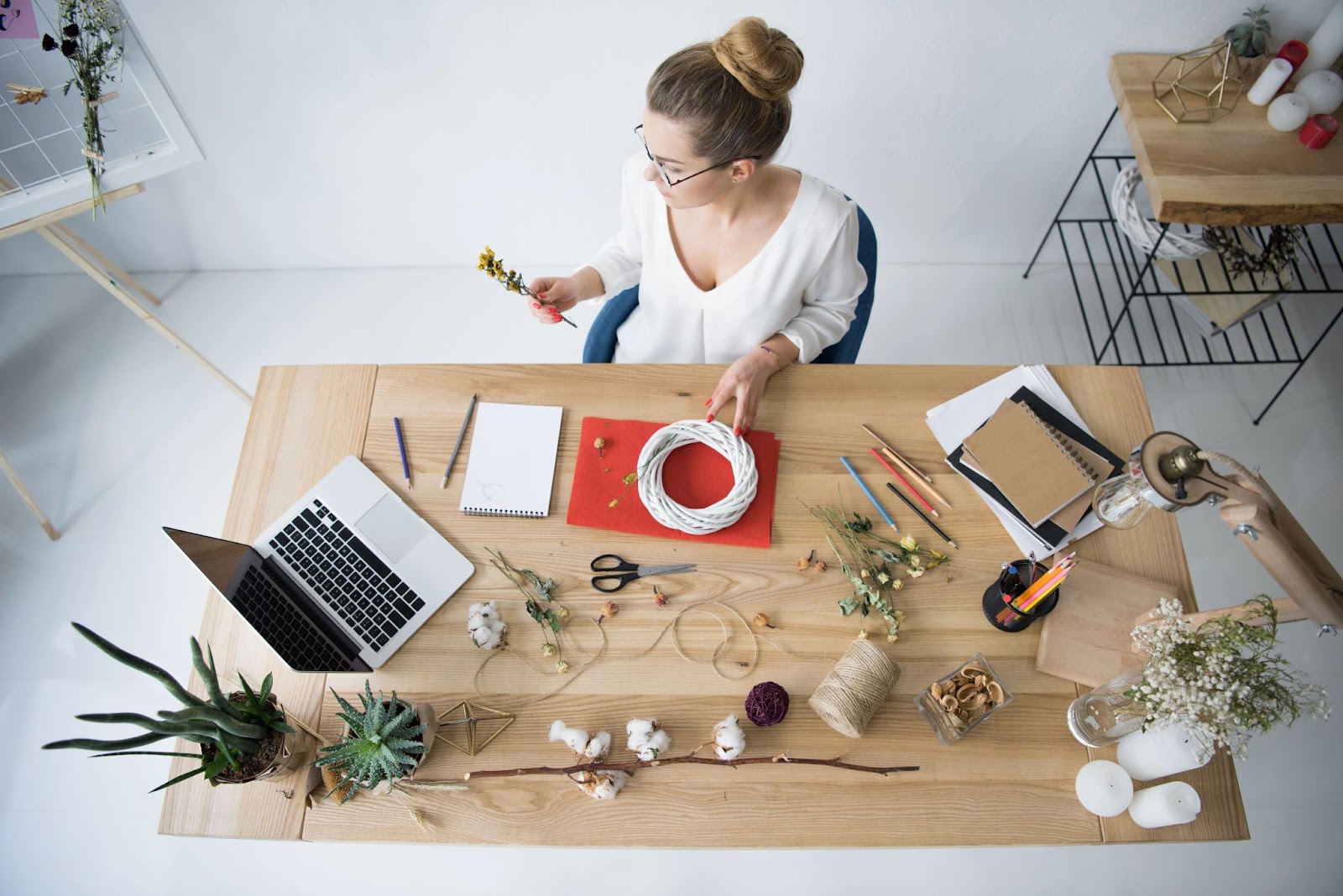 (A woman sits at a table with various crafting materials including wire, scissors, plants, and a laptop. She is holding a flower and looking at it thoughtfully as she tackles her latest DIY Craft Challenge project.)