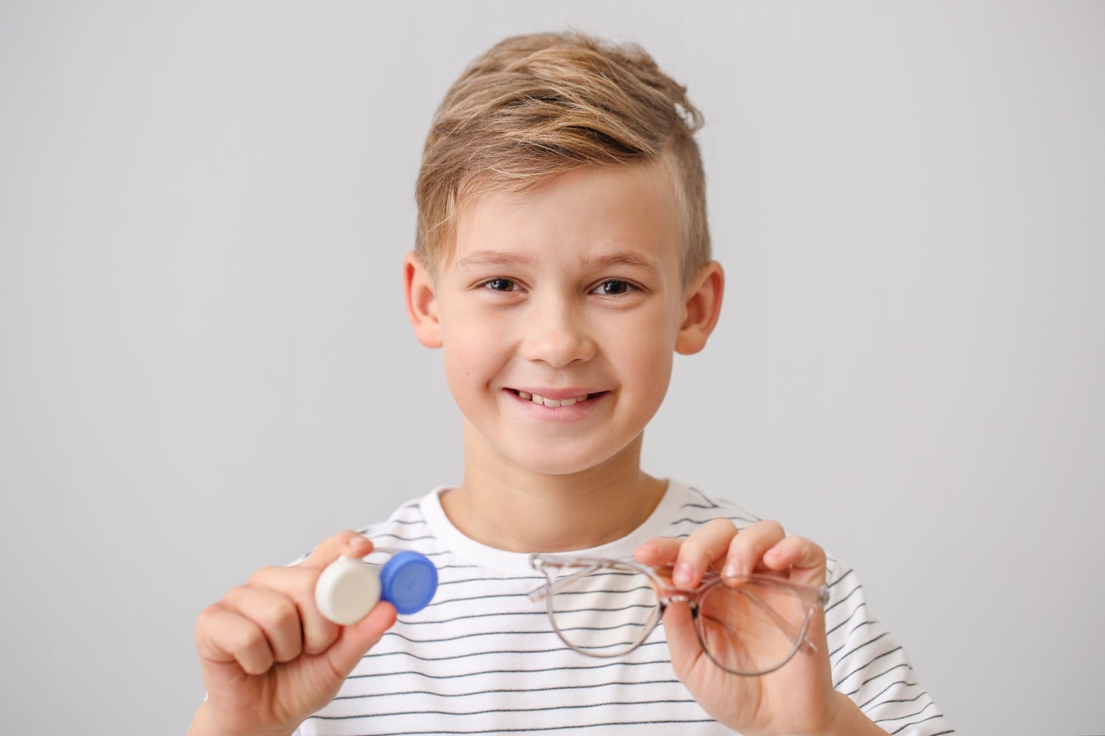 A young boy smiling and holing a contact lens case in his right hand and eyeglasses in his left hand.