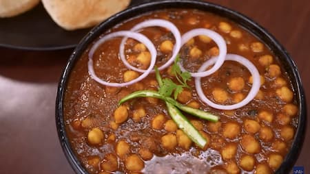 Chana masala garnished with chilies and onions, served hot in a bowl with puris on the side.