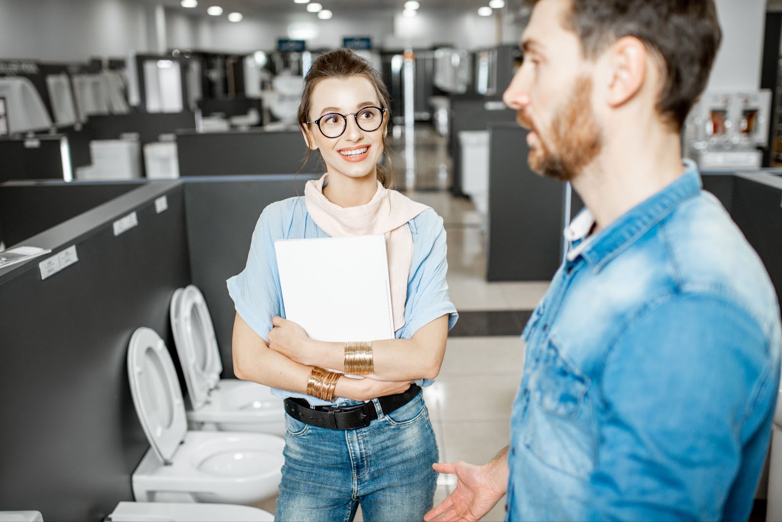 A bearded man in a denim long-sleeved polo shirt selects toilet ceramics in a shop, assisted by a bespectacled sales lady holding a folder. The background features a blurry array of assorted bathroom accessories.