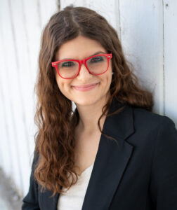 Portrait en buste de Rebel Weber, souriante, elle a des cheveux châtains longs et ondulés, de grandes lunettes rouges, et porte un tailleur.