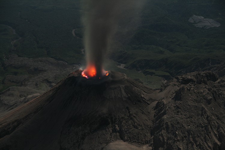 An aerial view of the Santiaguito volcano erupting at night. The volcano is spewing ash and lava, and it is surrounded by mountains.