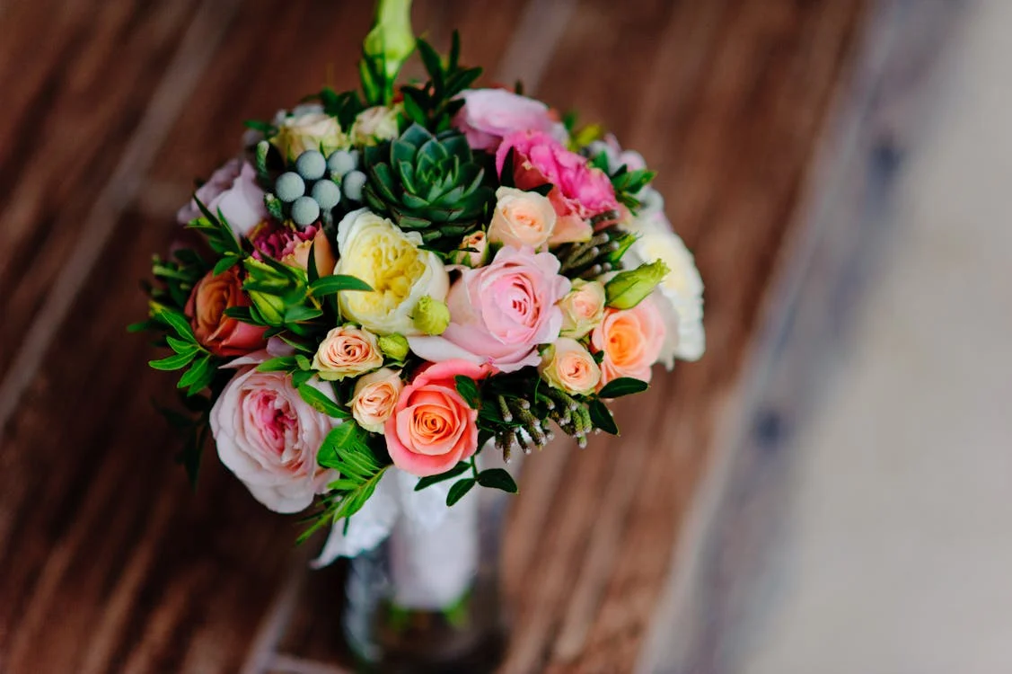 A vibrant bouquet of colorful flowers, including roses and succulents, arranged in a glass vase placed on a wooden table.