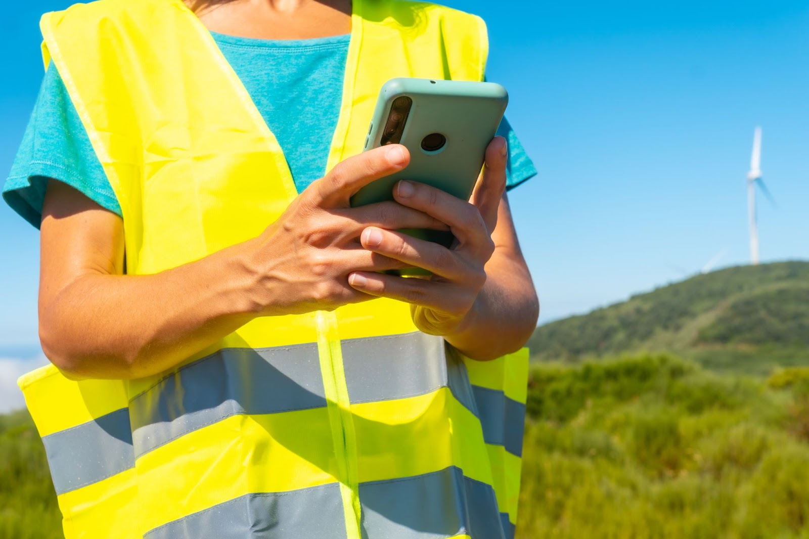 A half-body shot of a working woman in a neon yellow vest over a blue shirt, focusing on her hands holding a mobile phone while conducting a technical review at a wind farm.