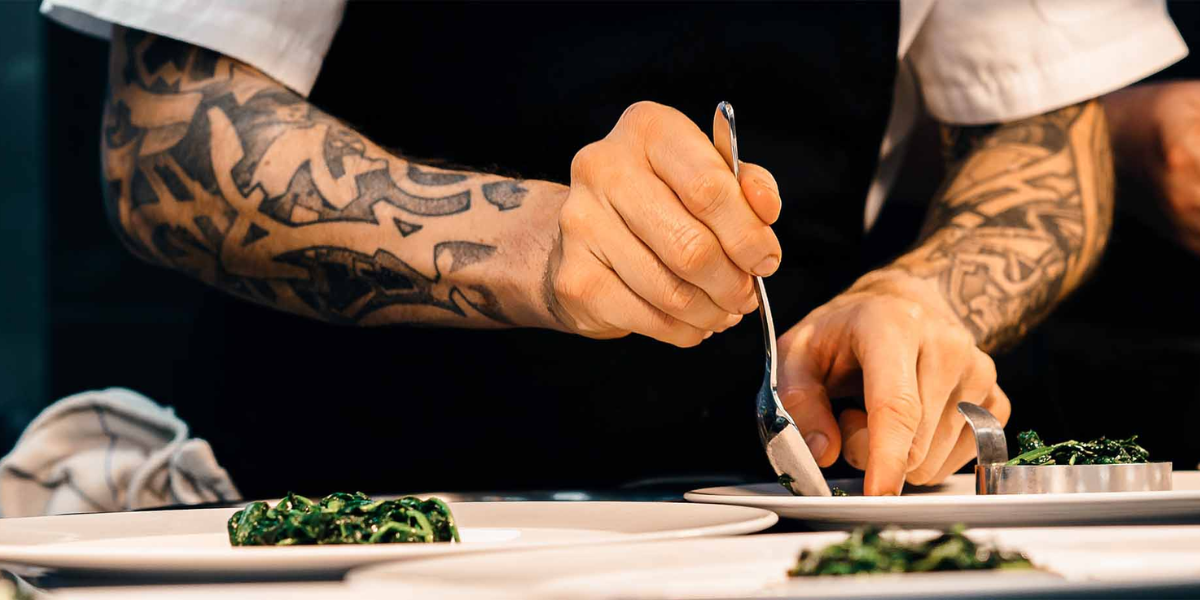 A tattooed chef carefully plating food in a professional kitchen.