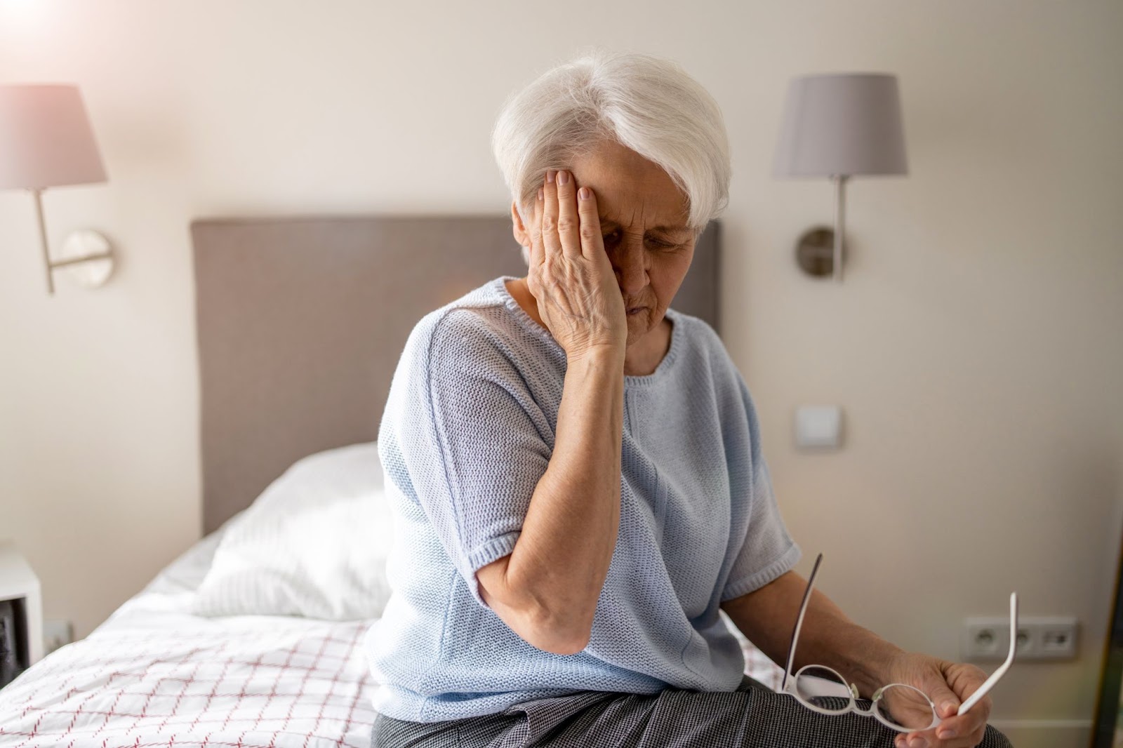 a senior woman sits on her bed holding her head, confused