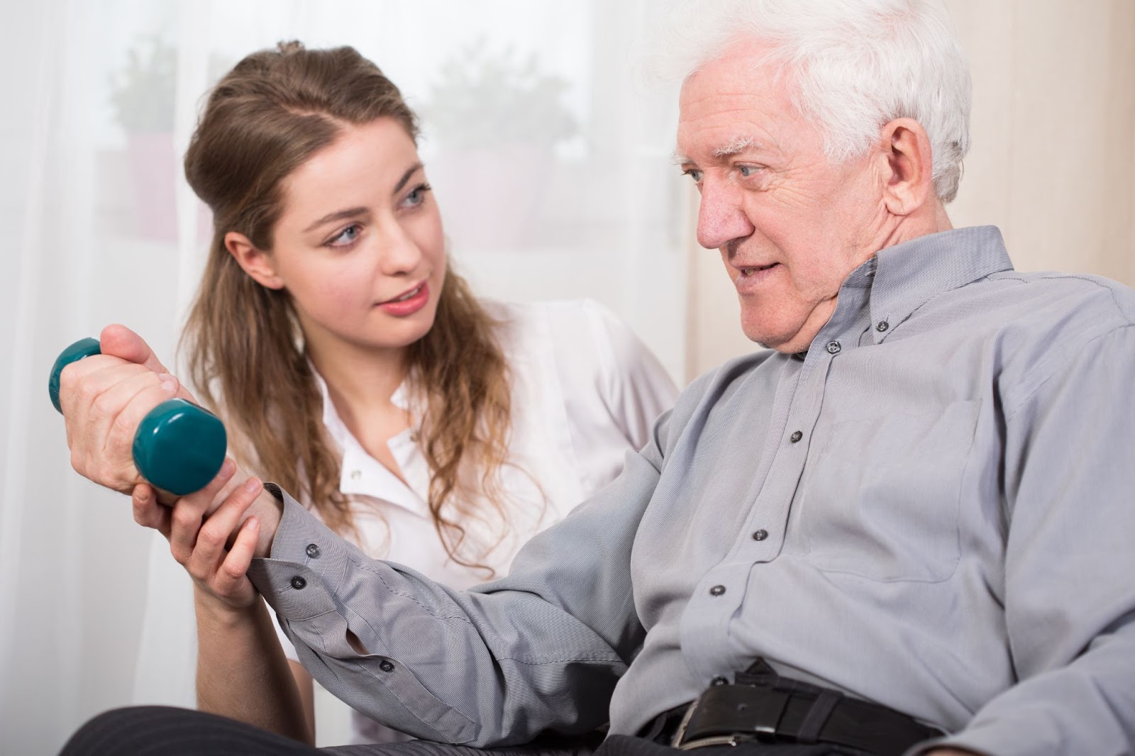 A female staff member helps an older adult sitting in a chair perform an exercise with a small dumbbell in his right hand.