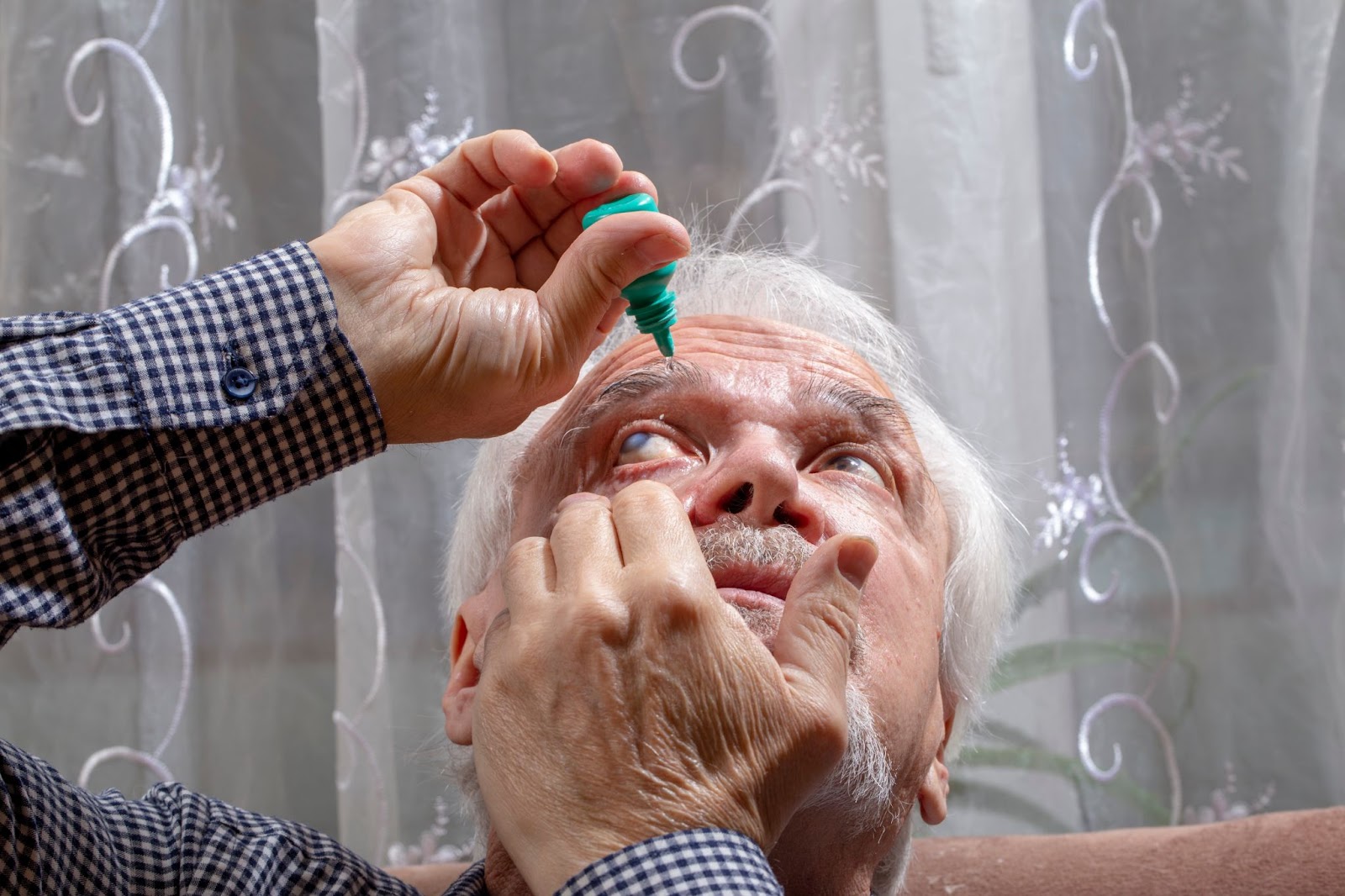 An older adult tilts his head back while pulling his right lower eyelid down with his left hand and dispensing eye drops with his right hand.