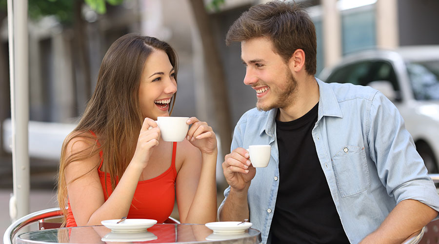 Brunette man and woman sitting next to each other and laughing while holding coffee cups.