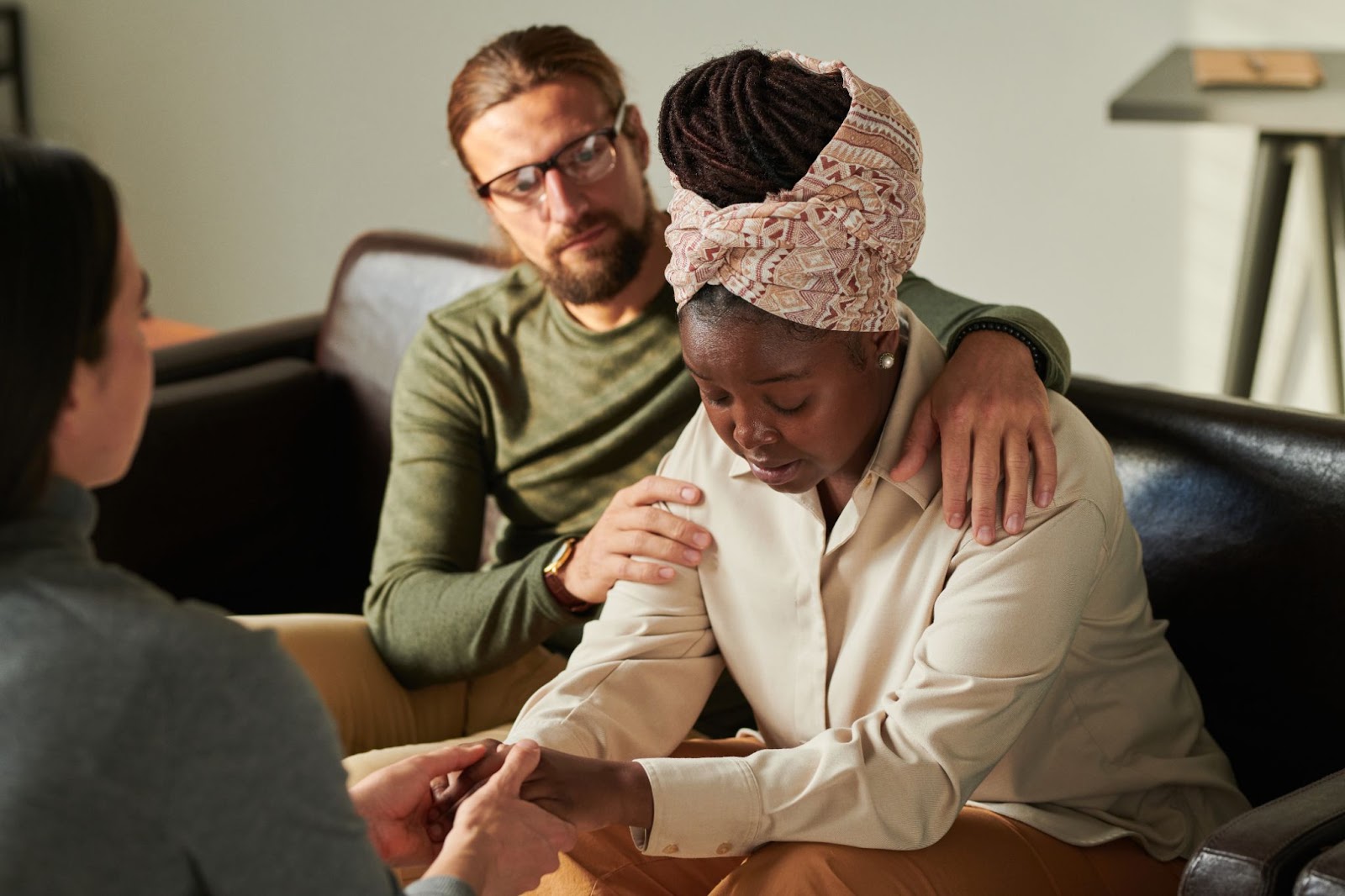 Woman and a man comforting a woman who has her eyes closed and is sitting on a couch. 
