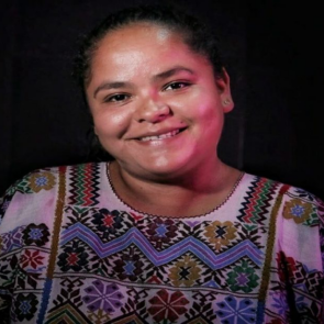 A close-up picture of Mexican indigenous WHRD Kenia Hernandez smiling. She is wearing a colourful embroidered shirt and standing in front of a black background. 