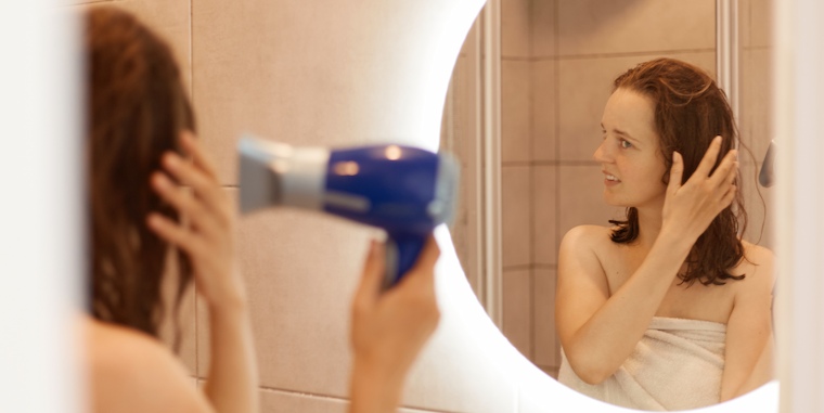 Imagem de uma mulher secando o cabelo com a ajuda de um secador elétrico. Ela está localizada dentro de um banheiro enquanto veste uma toalha.