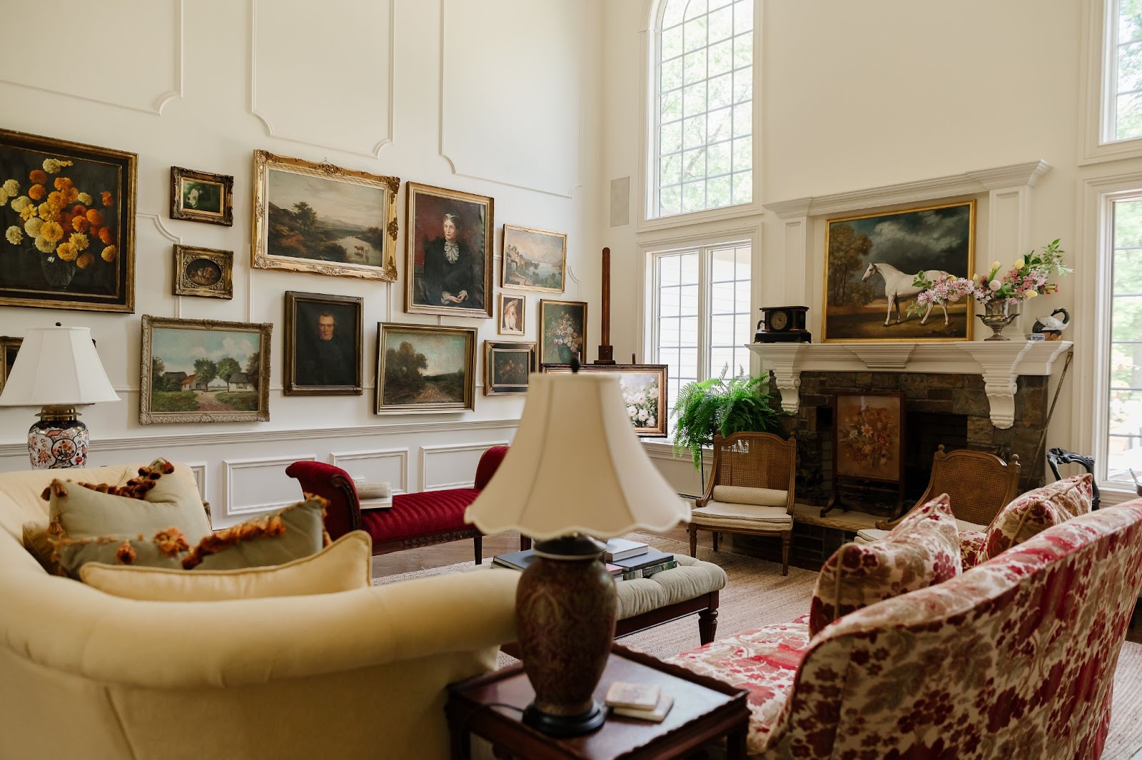 A high-ceiling and nicely appointed living room with palladian windows and warm white walls features a collection of beautifully framed wall art including portraits, equestrian scenes and landscapes.