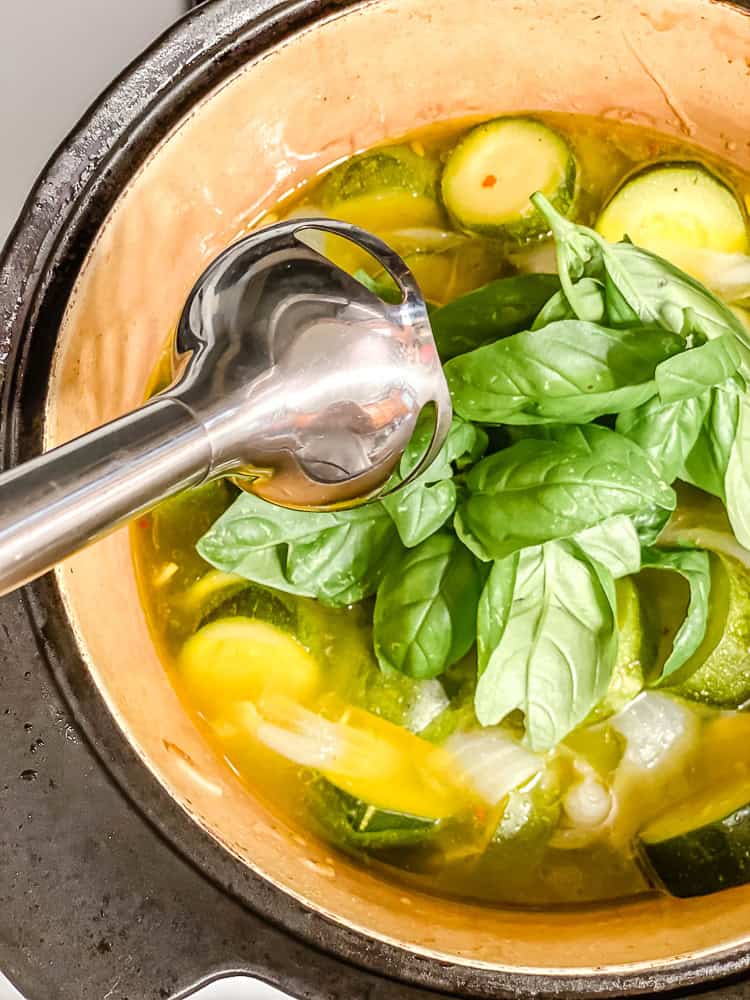 a metal immersion blender above a pot filled with basil leaves, water, and sliced zucchini.