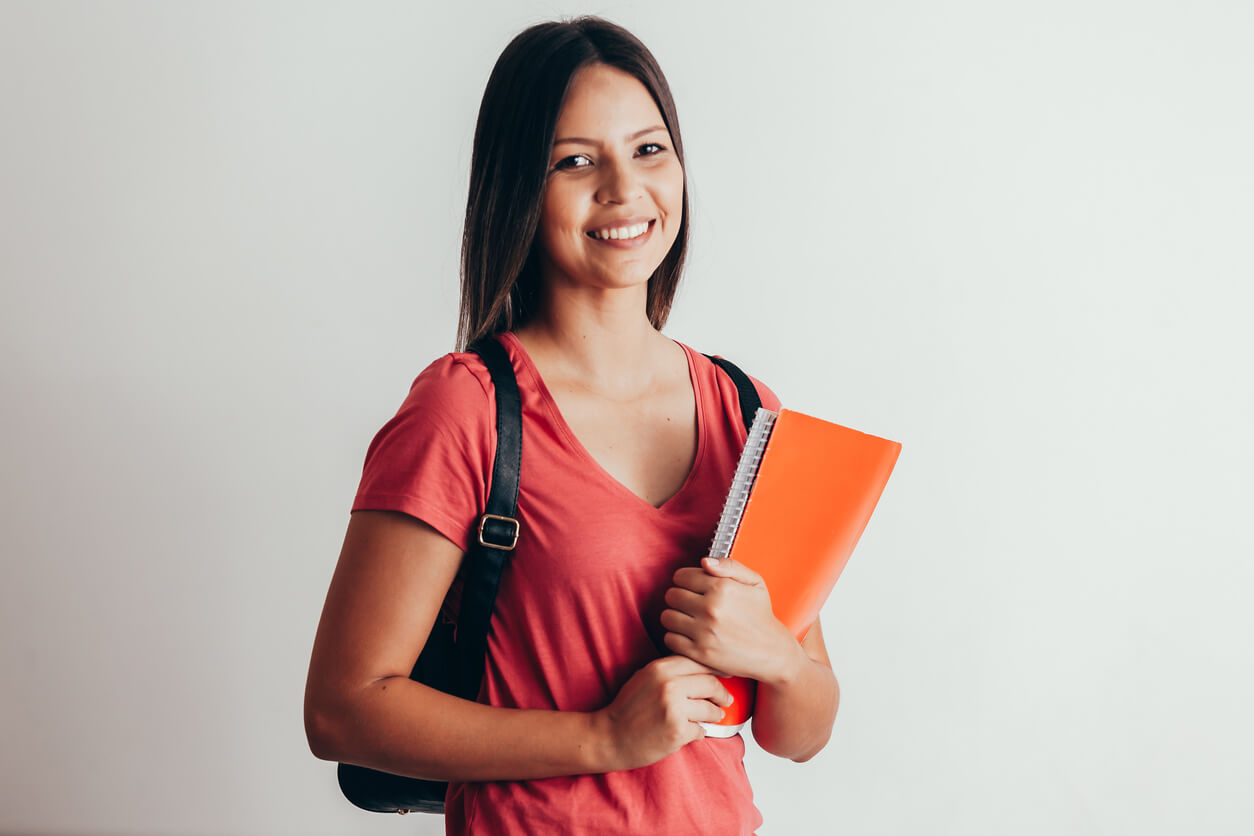 Imagem de uma estudante de blusa vermelha, segurando livros e sorrindo enquanto olha para a câmera.