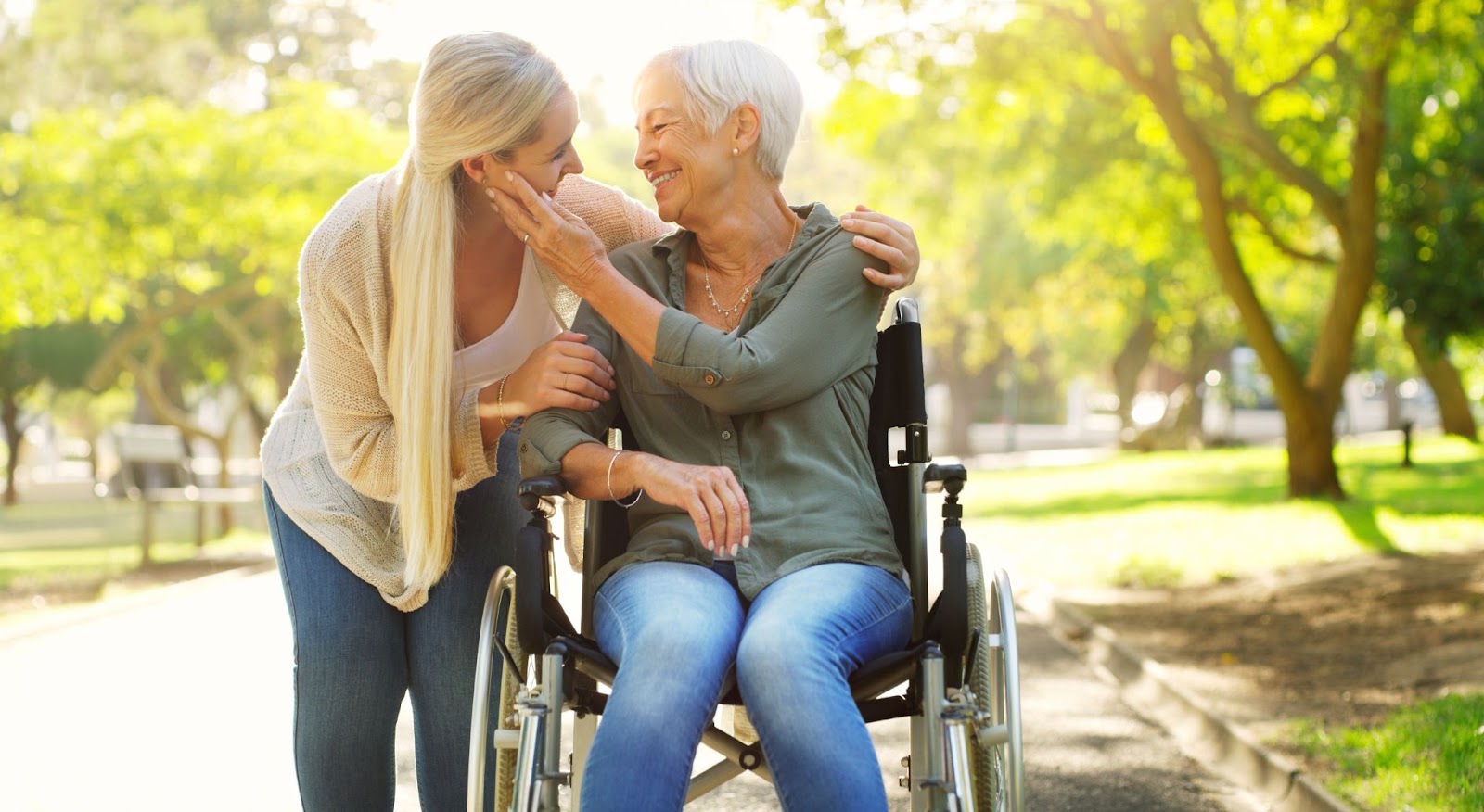 The daughter of an older adult woman in a wheelchair bends down to look at her mom while on a walk. They smile at each other.