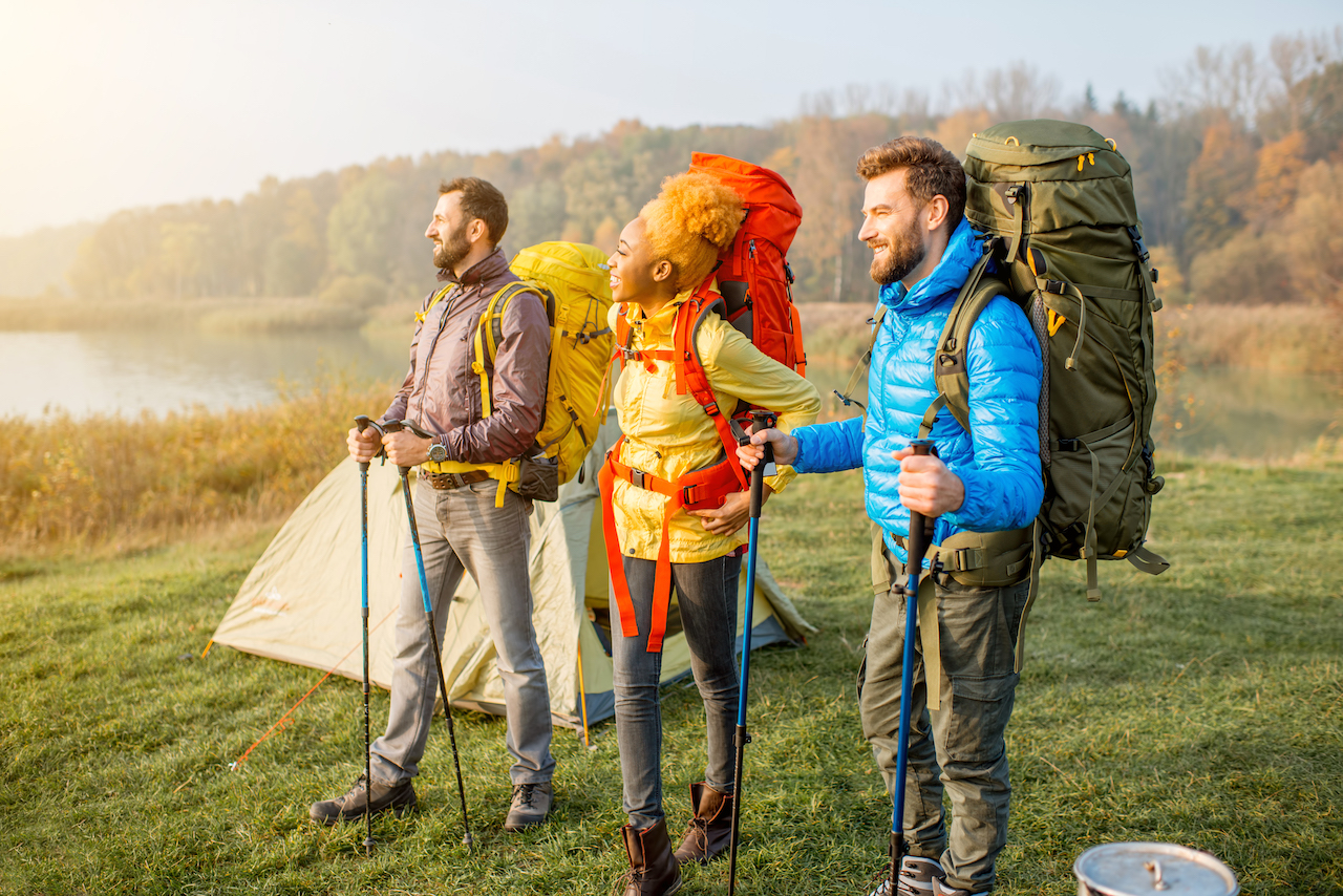 
Three people are dressed in appropriate camping gear while standing outdoors in front of a tent by a lake. They are each wearing sturdy hiking boots, colorful outdoor jackets, and large backpacks. The person on the left is in a purple jacket and gray pants, the person in the middle is in a yellow jacket and jeans, and the person on the right is in a blue jacket and olive-green pants. Each is holding a pair of trekking poles, and the scene is set against a backdrop of autumn foliage, suggesting it is a cool and crisp day. This image provides an excellent example of what to wear camping, including layers for warmth, durable footwear, and essential gear for hiking and backpacking.