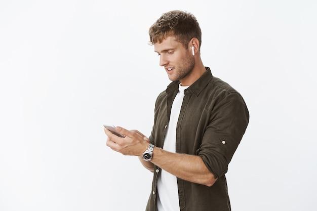 Waist-up shot of good-looking european man getting outside wearing wireless earphones holding smartphone looking at cellphone screen, listening to music