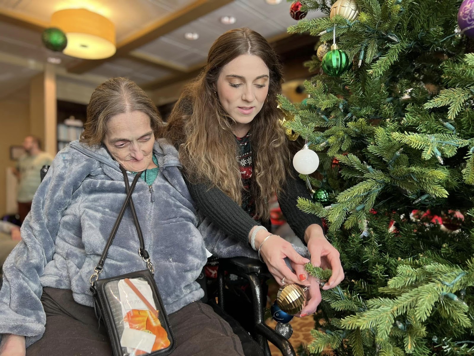 A memory care worker helping an elderly woman in a wheelchair place ornaments on a Christmas tree