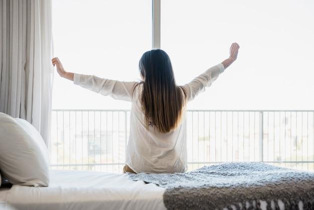 Free photo rear view of a woman sitting on bed stretching her arms