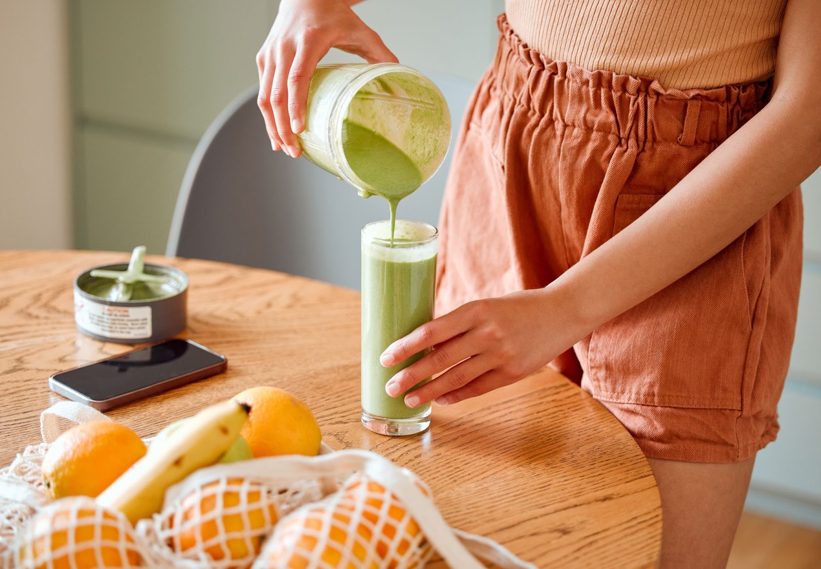 Close-up of a girl's hands pouring a green smoothie from a magic bullet into a glass on a kitchen table.