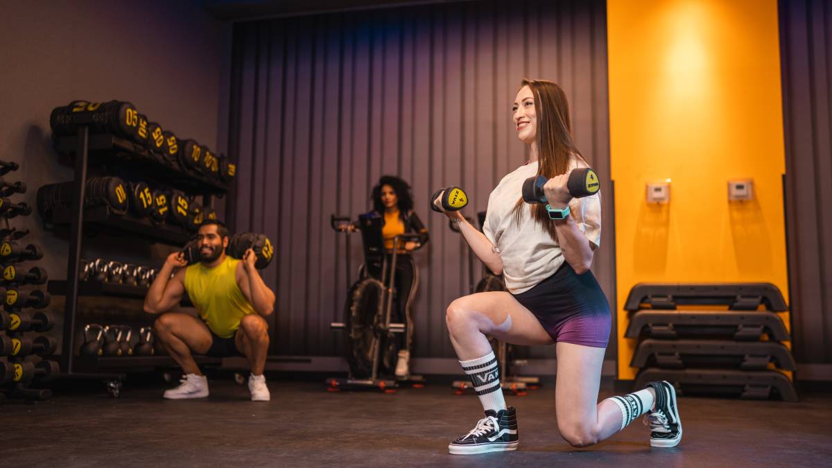 mujer realizando un desplante e un gimnasio Smart Fit, con una mancuerna en cada mano y una sonrisa en el rostro