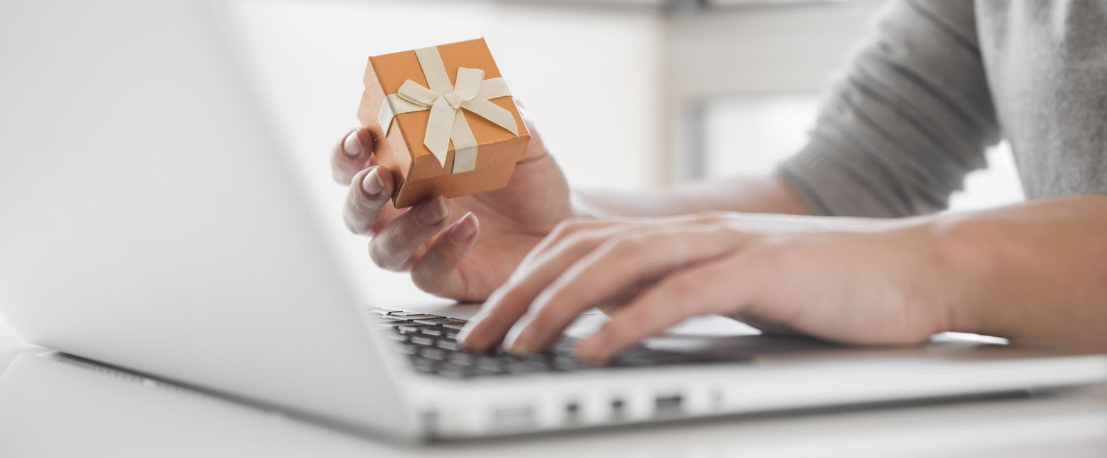 Pessoa segurando uma pequena caixa de presente com um laço enquanto digita em um laptop, simbolizando compras online e programas de fidelidade.  mercado de fidelização