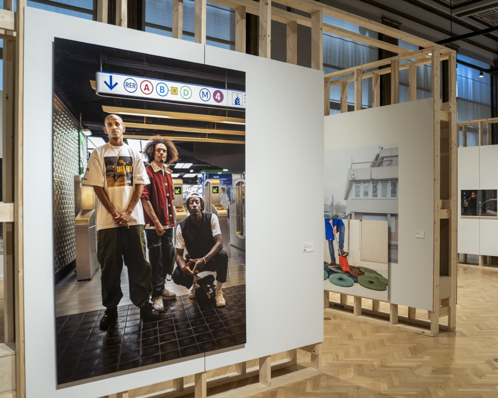 Image: Installation view: A photo from Tonika Lewis Johnson’s Belonging series featuring three young Black men outside the turnstiles of a Paris metro station. Two of the men are standing, and the third is squatting. All three are looking directly into the camera. In the background, other photographs from the installation are partially visible. Courtesy of Villa Albertine.