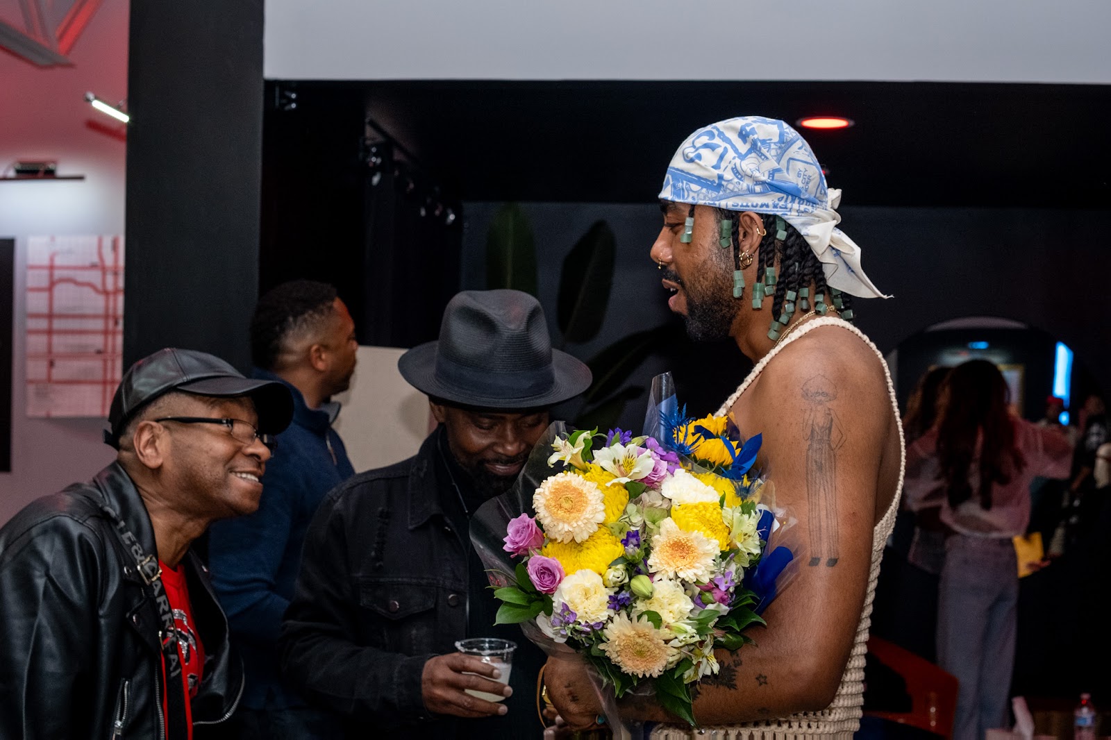 Image: Tisha Taylor and Nasir Montalvo interacting at the {B/qKC}: The Volume_2 Archive Launch Party, Montalvo holds flowers and wears a blue-and-white bandana on their head. Taylor smiles, wearing a black leather ball cap and a black leather jacket. Between them is another person wearing a black broad-rimmed hat. Behind them, are people looking further back in the room. Photo by Gabriella Salinas.