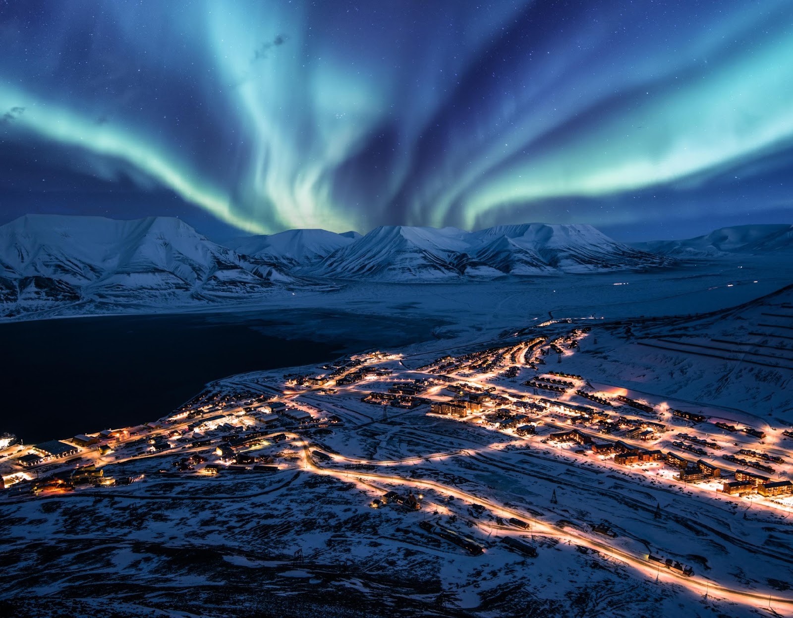 A scenic view of snowcapped mountains illuminated by ribbons of aurora. City lights flow like ribbons from below the mountains.