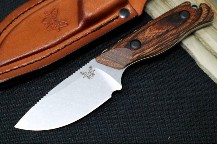 An image featuring the Benchmade 15017 Hidden Canyon Hunter on a black and wooden background. Next to the knife is a fitted leather sheath with the Benchmade butterfly logo embossed on it.