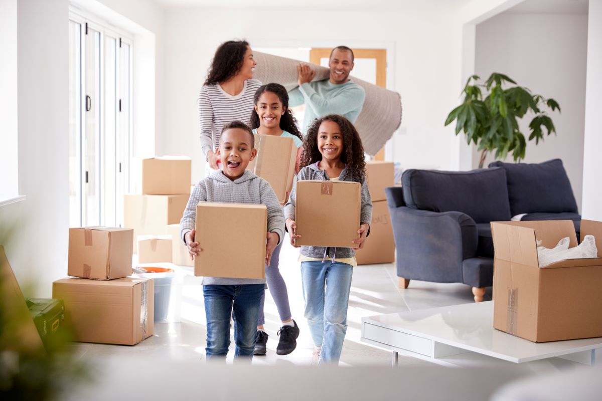 Smiling family carries boxes and a rug into their new home.