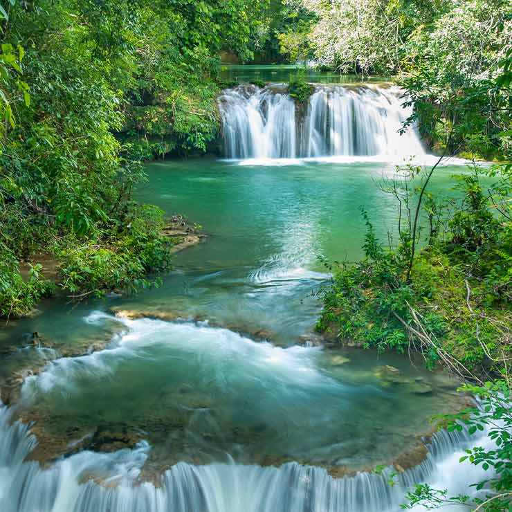 Cachoeira cercada por vegetação densa, com águas cristalinas fluindo para uma lagoa verde-esmeralda tranquila