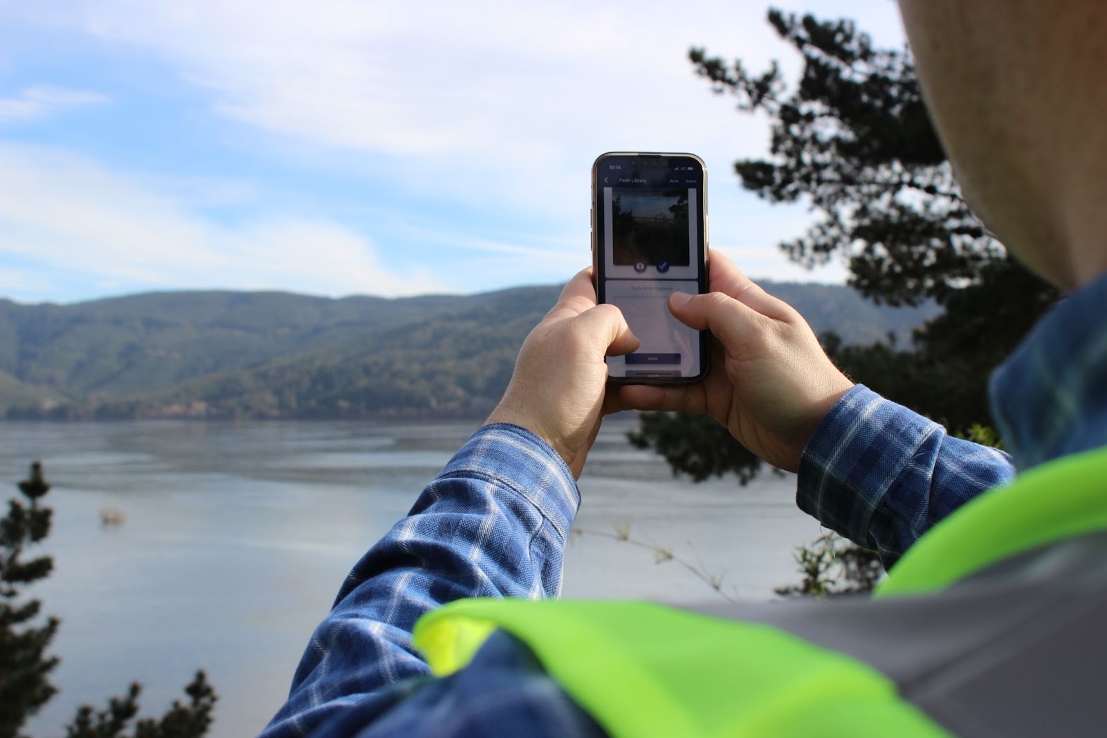 Someone holding a phone with both hands with water and mountains in the background. 