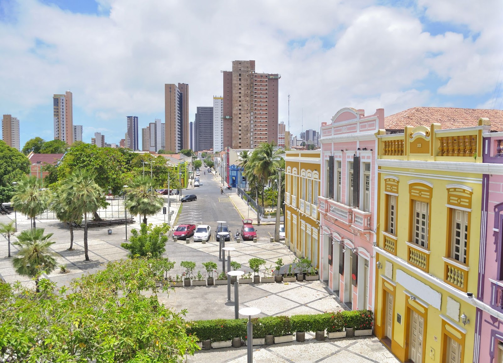 Casas da Rua Dragão do Mar, na parte central de Fortaleza. As antigas construções estreitas em estilo holandês estão enfileiradas de frente para uma praça e têm fachadas coloridas bem preservadas. Ao fundo, aparecem prédios mais modernos e uma avenida.