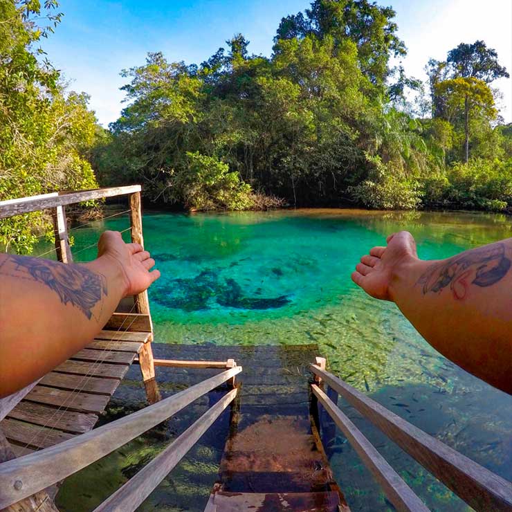 Pessoa com braços tatuados estendidos, de frente para uma lagoa de águas cristalinas e escadaria de madeira, rodeada por vegetação densa