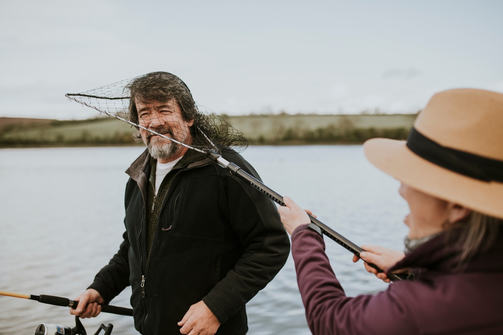 An older adult man and woman enjoy a playful moment while fishing as the woman captures the man's head in a fish net.