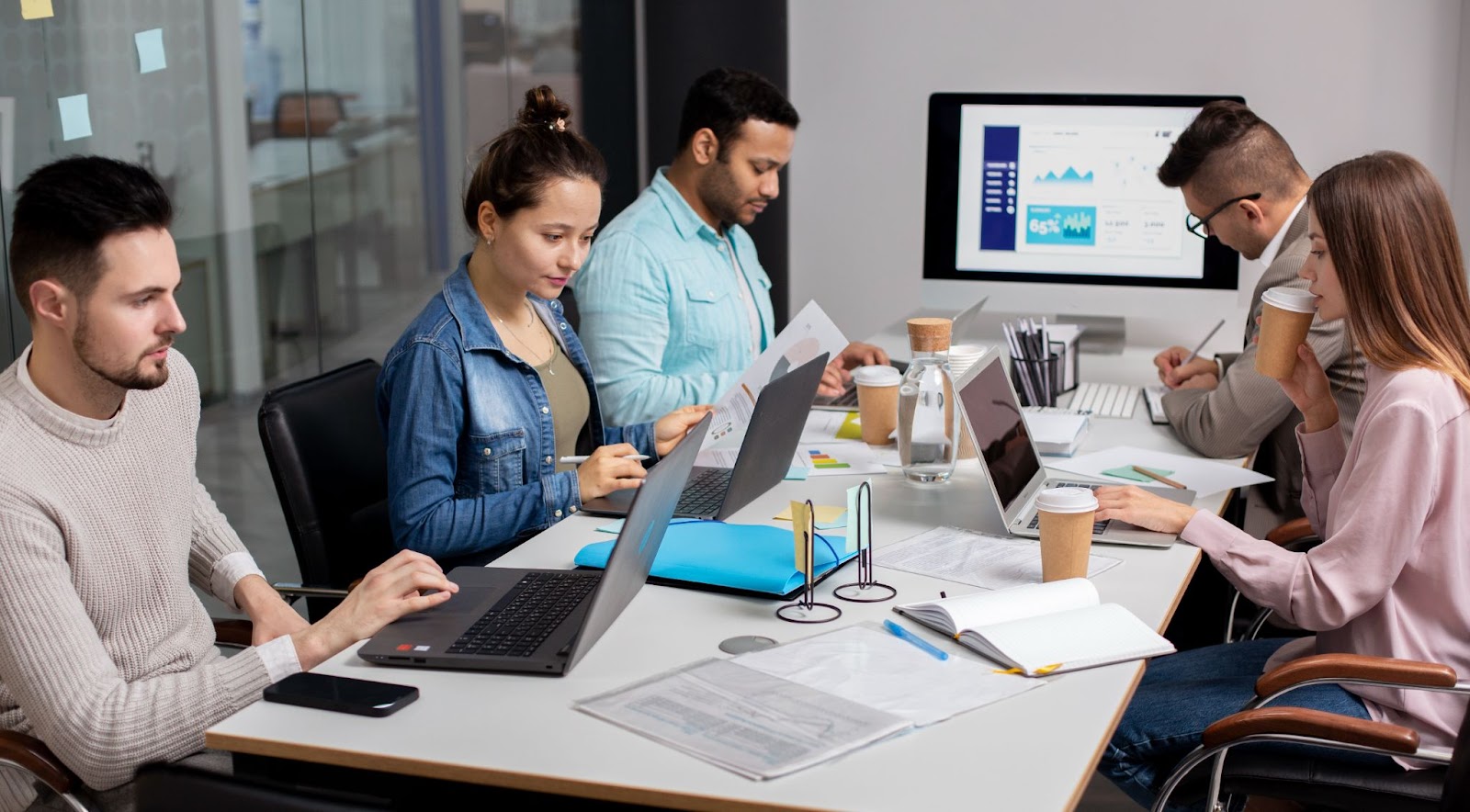 A group of five people are working in an office setting in the best digital marketing agency in Kochi. They are seated around a table, each focused on their laptops and paperwork. The office appears modern and well-equipped, with a large monitor displaying charts and graphs in the background. Various documents, notebooks, and coffee cups are scattered on the table, indicating a productive work session.