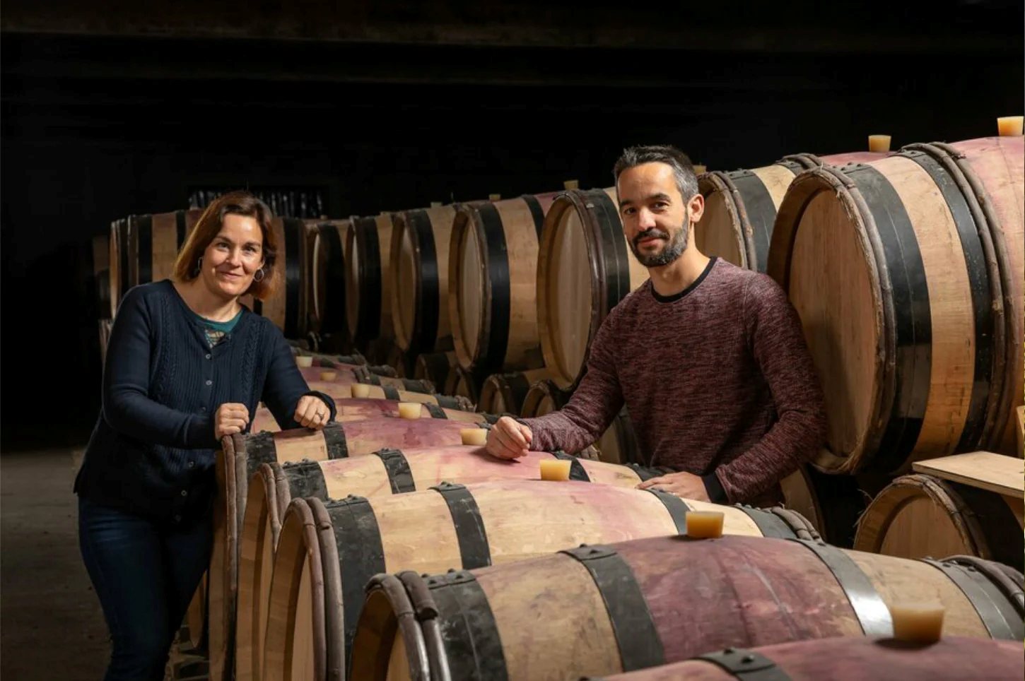 A person and person standing next to a stack of wooden barrels

Description automatically generated