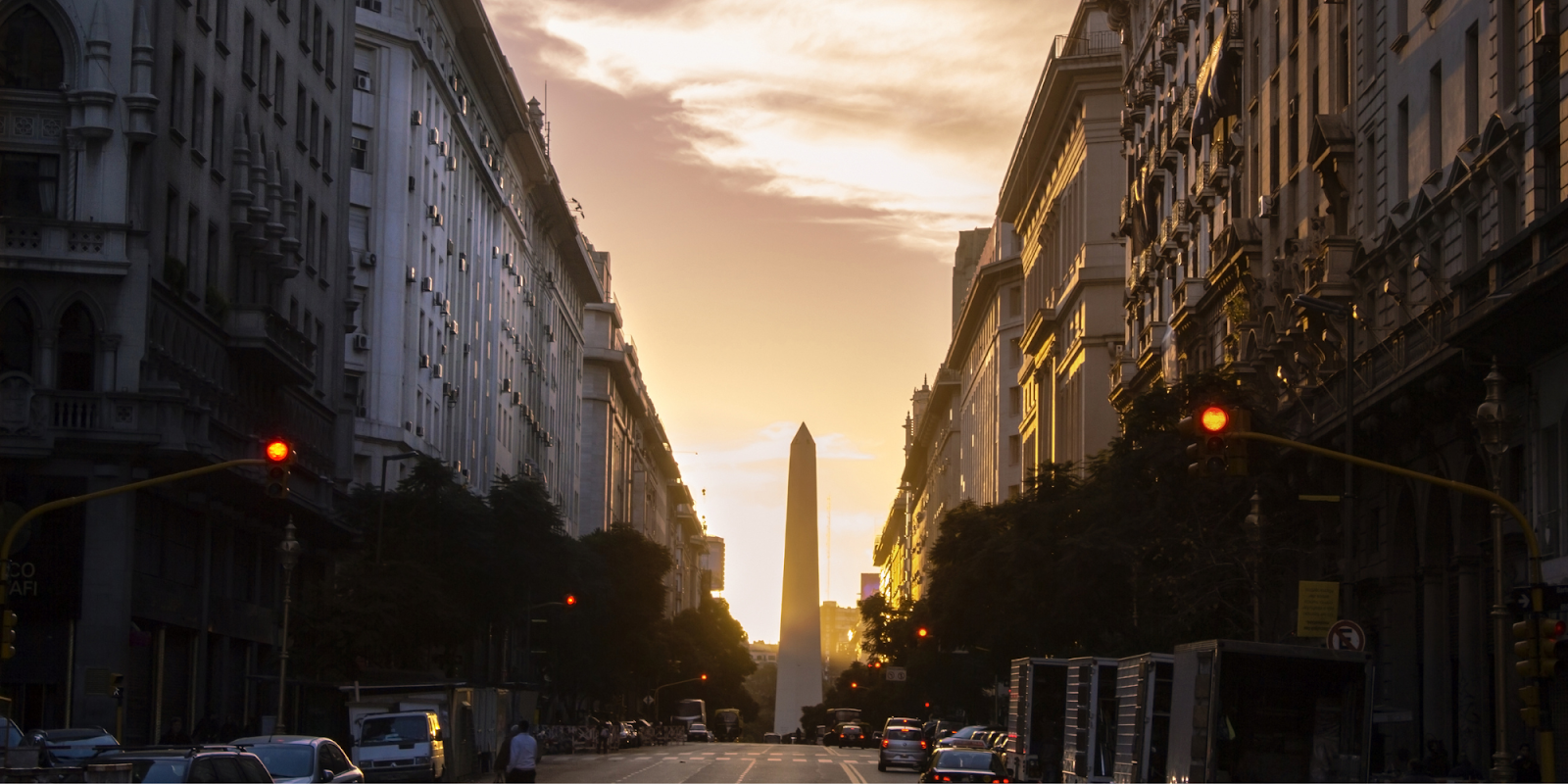 A captivating view of Buenos Aires, Argentina, at sunset, with the iconic Obelisk of Buenos Aires prominently featured in the distance. The street is lined with tall buildings on both sides, casting shadows as the golden light of the setting sun creates a warm glow. Traffic lights and vehicles add to the urban atmosphere, highlighting the city's bustling yet serene vibe as day turns to evening.