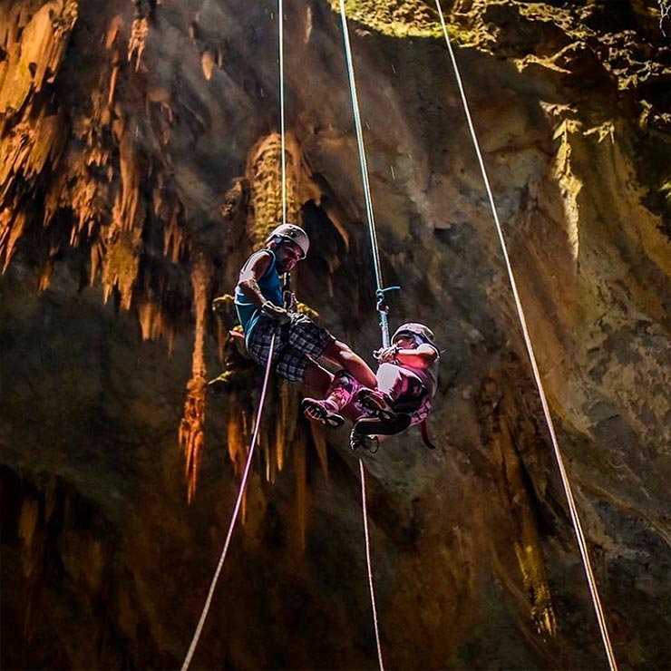 Duas pessoas usando capacetes e equipamentos de segurança, praticando rapel em uma caverna com iluminação dramática