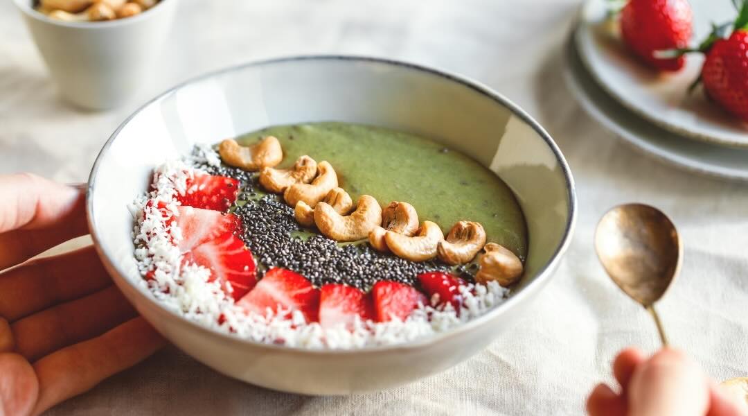 A healthy smoothie bowl topped with fresh strawberries, cashews, chia seeds, and shredded coconut, with hands holding the bowl and a spoon nearby.