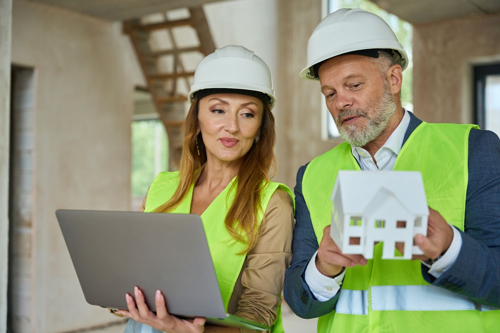 A woman wearing a yellow vest and a white hard hat is holding a laptop while a man next to her, wearing a white hard hat and yellow vest, is showing the woman a replica of a house.