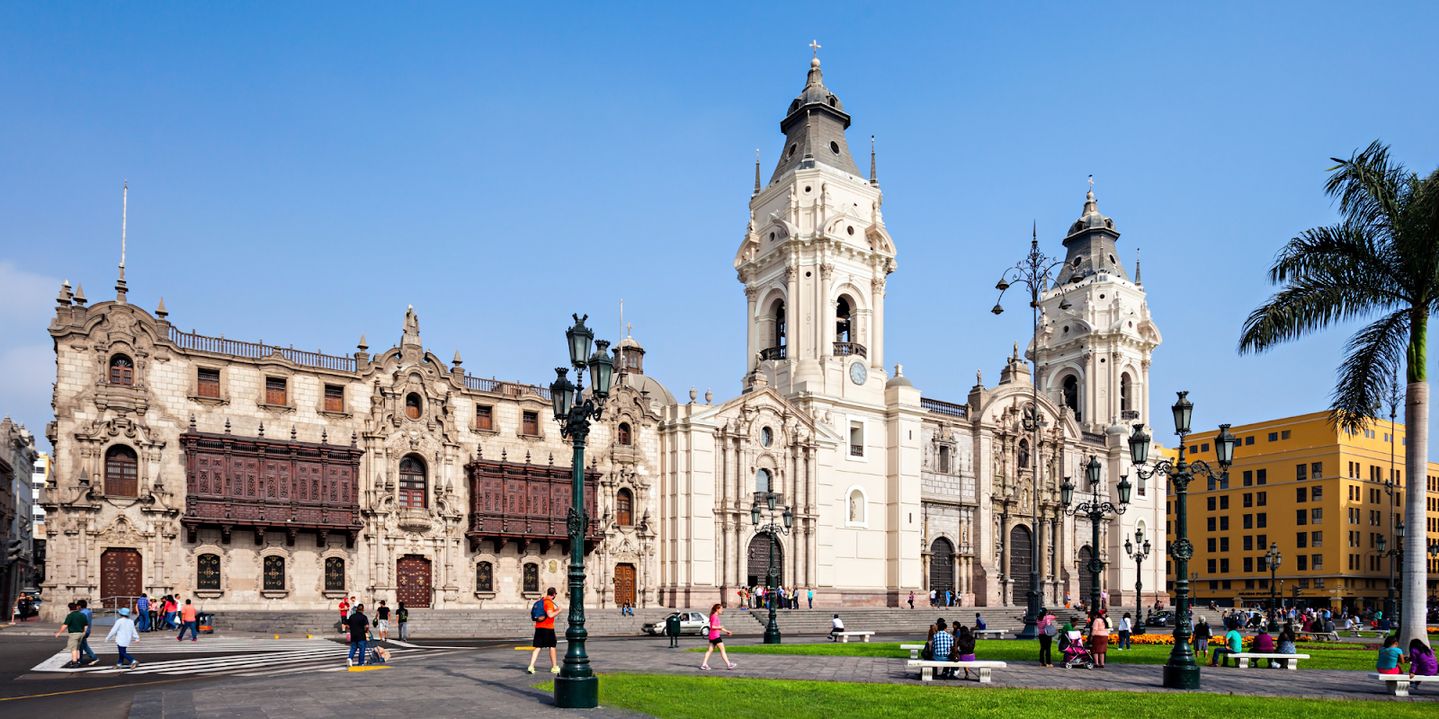 A bright and sunny day at Plaza Mayor in Lima, Peru, featuring the beautiful historic buildings of the Archbishop's Palace and the Cathedral of Lima. People are strolling around the square, enjoying the pleasant weather. The lush green grass, palm trees, and classic street lamps add to the charm of this central and significant location in Lima. The intricate architectural details of the buildings are highlighted against the clear blue sky.