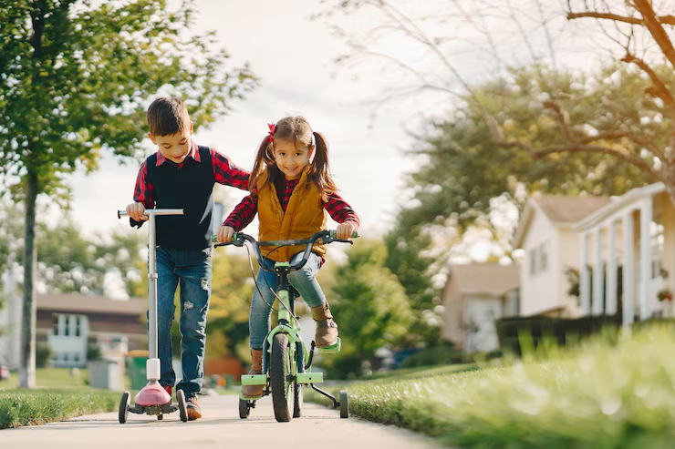 A boy riding a scooter while helping his sister ride a bike