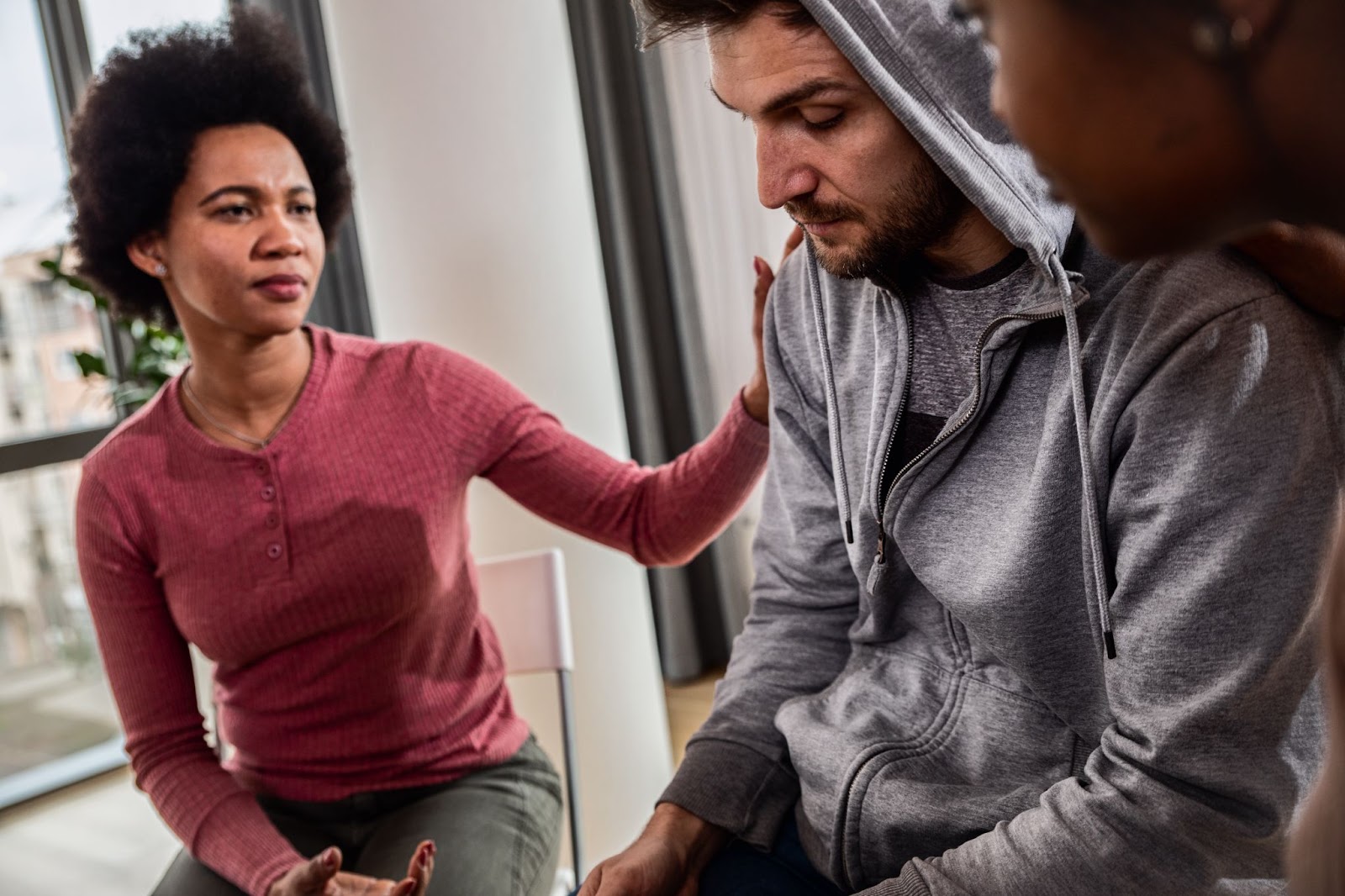 A man wearing a gray sweatshirt in a group therapy setting looks sad and distressed while others console him.