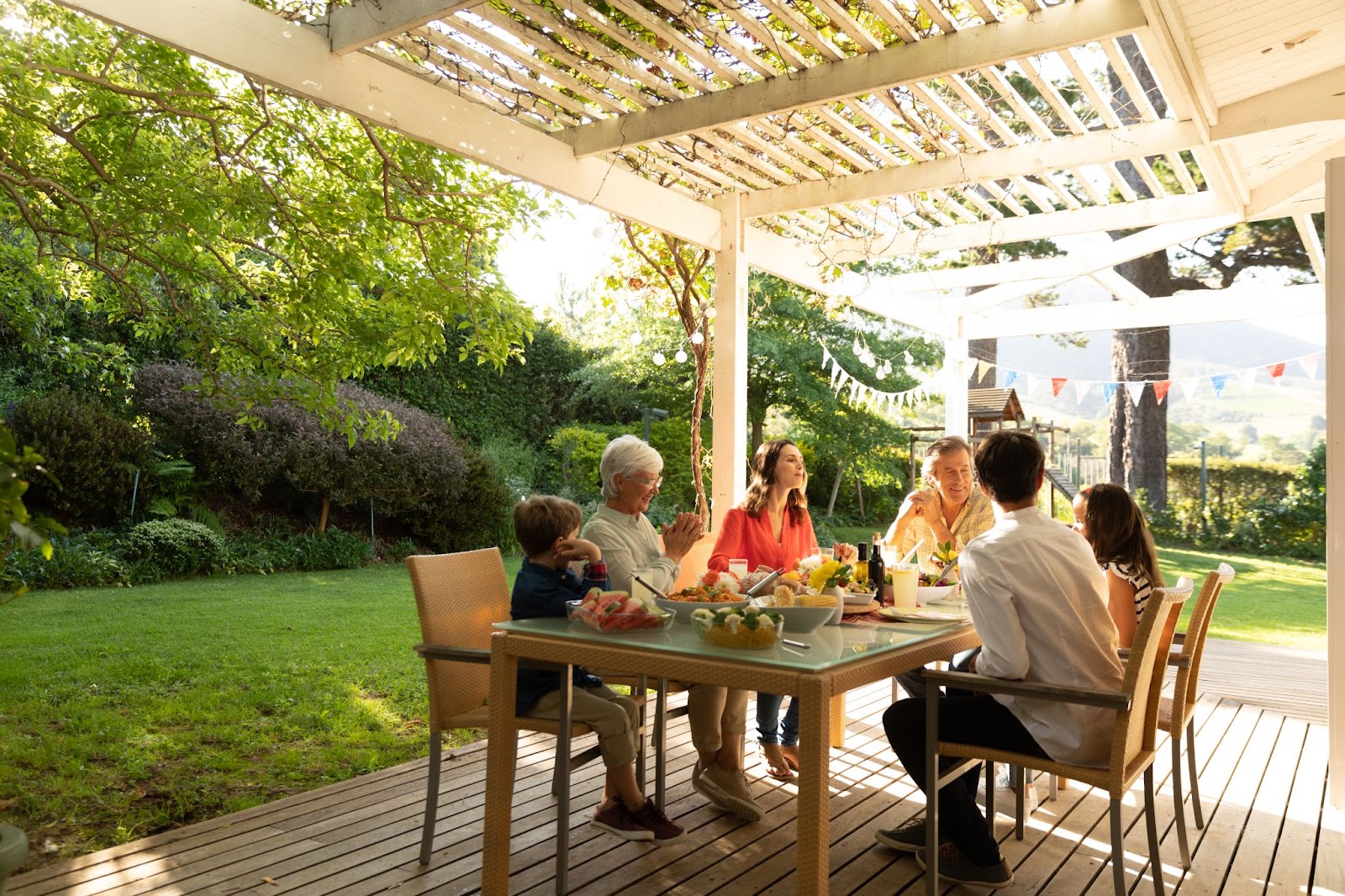 A family enjoys a meal on a covered patio, with a lush garden as a backdrop on a sunny summer day.