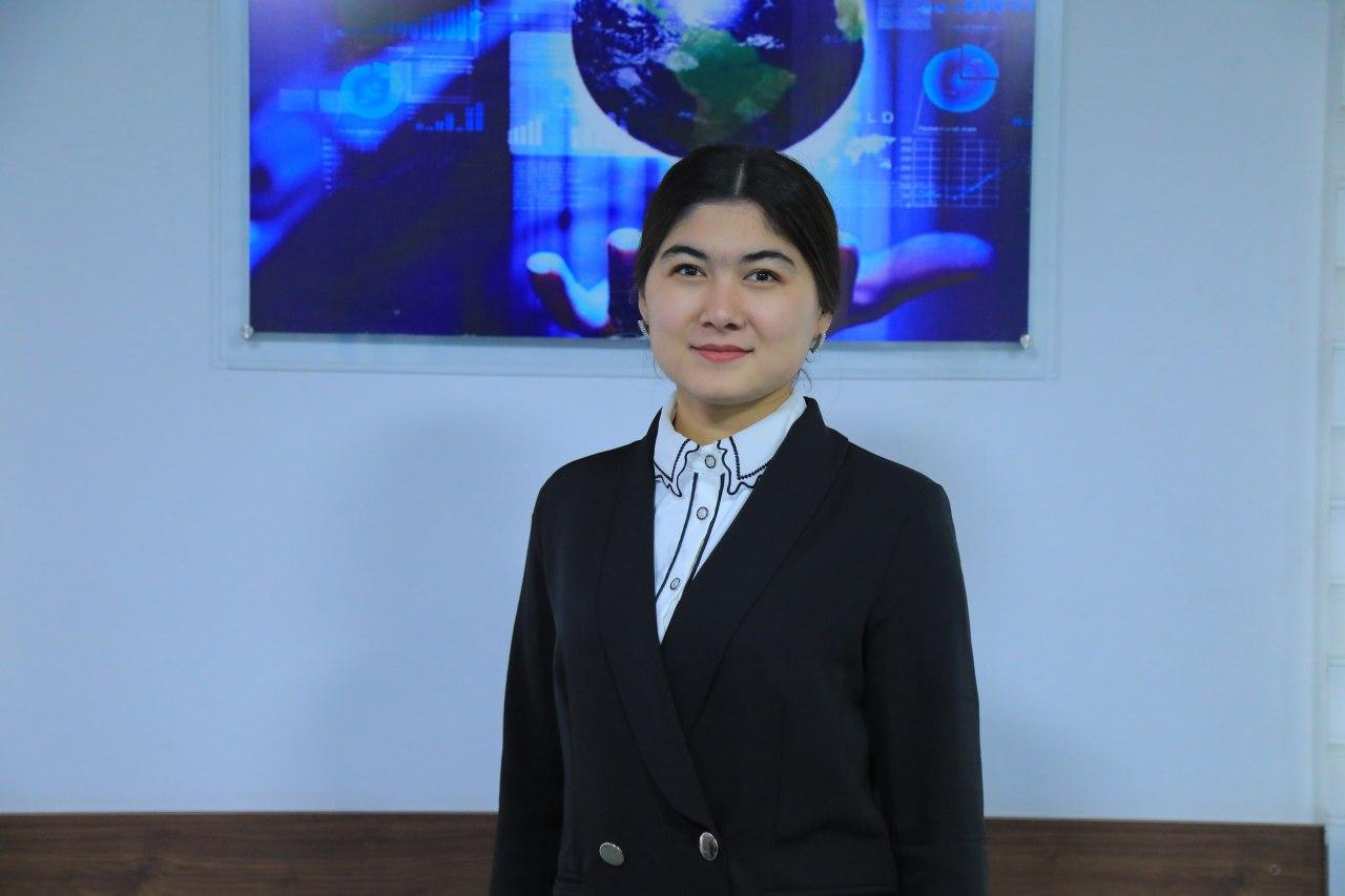 Central Asian woman in a black coat and white blouse standing in front of a wall with a picture of a globe on it. 